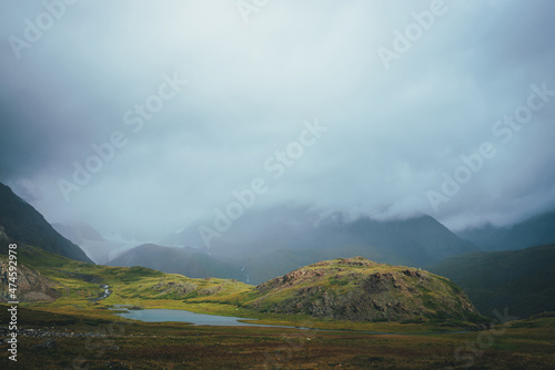Dramatic alpine landscape with beautiful mountain lake in green valley among great rocks and high mountains in overcast weather. Awesome view to lake and glacier in low clouds. Atmospheric scenery.