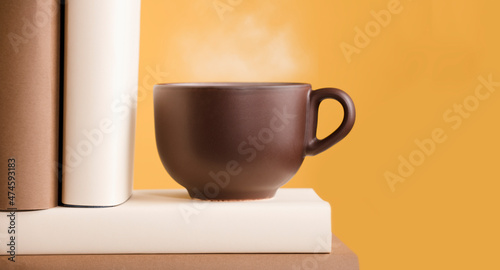 Cup of coffee or tea with hot steam on book on shelf orange yellow background