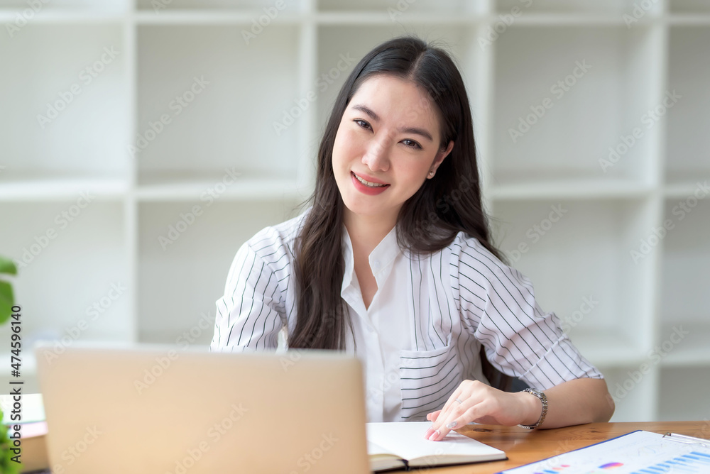 Smiling beautiful young Asian businesswoman sitting with laptop and computer while doing some paperwork at the office. Looking at camera. Smiling beautiful young Asian businesswoman sitting with laptop and computer while doing some paperwork at the office. Looking at camera.