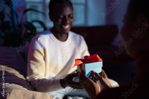 Young african woman giving gift box to boyfriend, congratulating husband with birthday, happy black man on sofa receiving unexpected present on Valentines day from wife at home. Selective focus