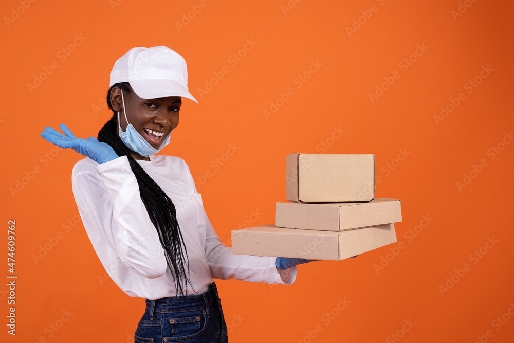 delivery, pandemic, helthcare and people concept - a happy African-American female courier in a pulled down protection mask and gloves holding boxes in the palm on orange background with copy space delivery, pandemic, helthcare and people concept - a happy African-American female courier in a pulled down protection mask and gloves holding boxes in the palm on orange background with copy space