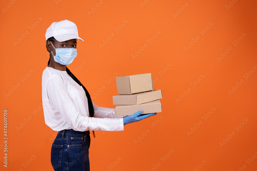 delivery, pandemic, helthcare and people concept - a confident African-American female courier standing sideways in a protective mask holding boxes on palms on orange background with copy space delivery, pandemic, helthcare and people concept - a confident African-American female courier standing sideways in a protective mask holding boxes on palms on orange background with copy space