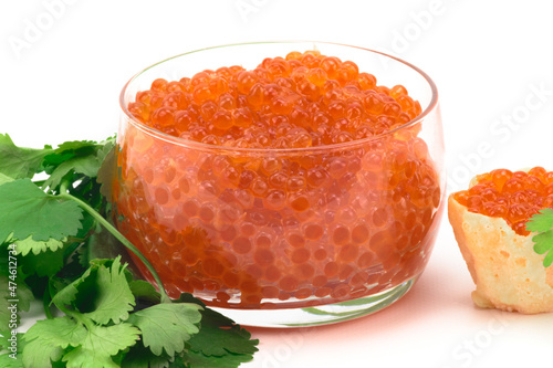 A large glass bowl filled with red caviar, next to it there is a bunch of parsley, and on the other side there is a tartlet with caviar