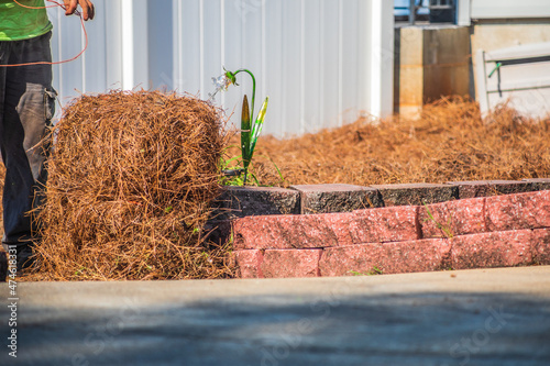 Bales of pinestraw laying beside a driveway flowerbed with brick edging and a white fence while a landscaper works in the background
