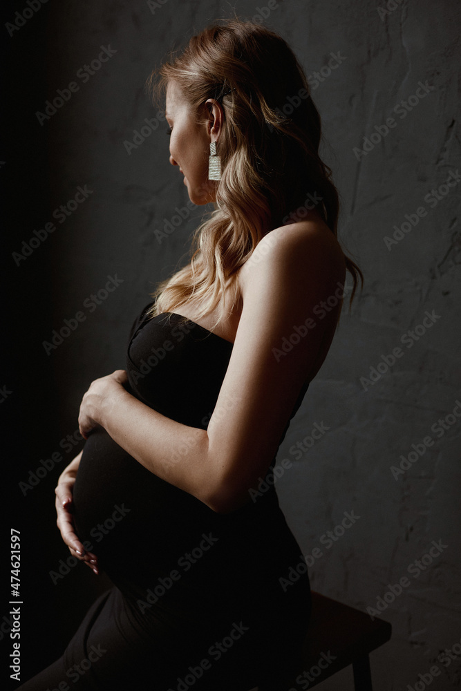 Young pregnant woman with long blond hair wearing black dress keeping her hands o her belly and looking aside Young pregnant woman with long blond hair wearing black dress keeping her hands o her belly and looking aside