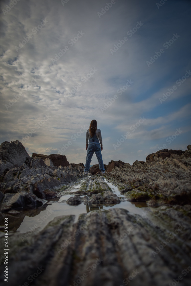 girl in denim overalls, looking towards the sea on some stones