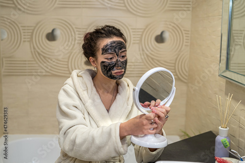 the girl puts a black mask on her face made of black clay and corrects it to the mirror.