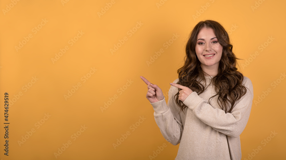 A young, happy and attractive Caucasian brunette girl with wavy hair in a casual jumper pointing her fingers at an empty advertising space on an orange studio background. A young, happy and attractive Caucasian brunette girl with wavy hair in a casual jumper pointing her fingers at an empty advertising space on an orange studio background.