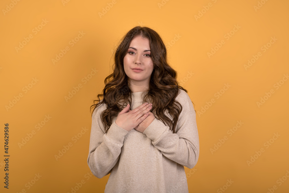 A beautiful and attractive Caucasian brunette girl in a casual jumper showing gratitude while holding her hands on her chest on an orange studio background. A beautiful and attractive Caucasian brunette girl in a casual jumper showing gratitude while holding her hands on her chest on an orange studio background.
