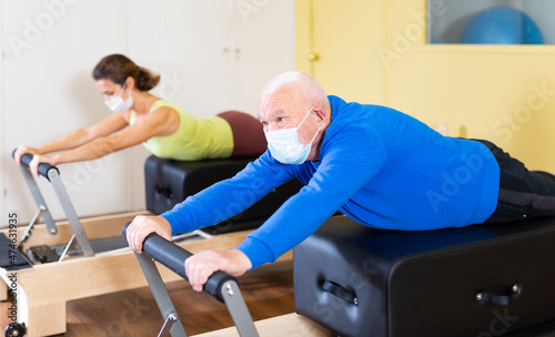 Focused senior man wearing medical mask to protect against viral infections practicing pilates stretching exercises on reformer as part of remedial gymnastics.. photo