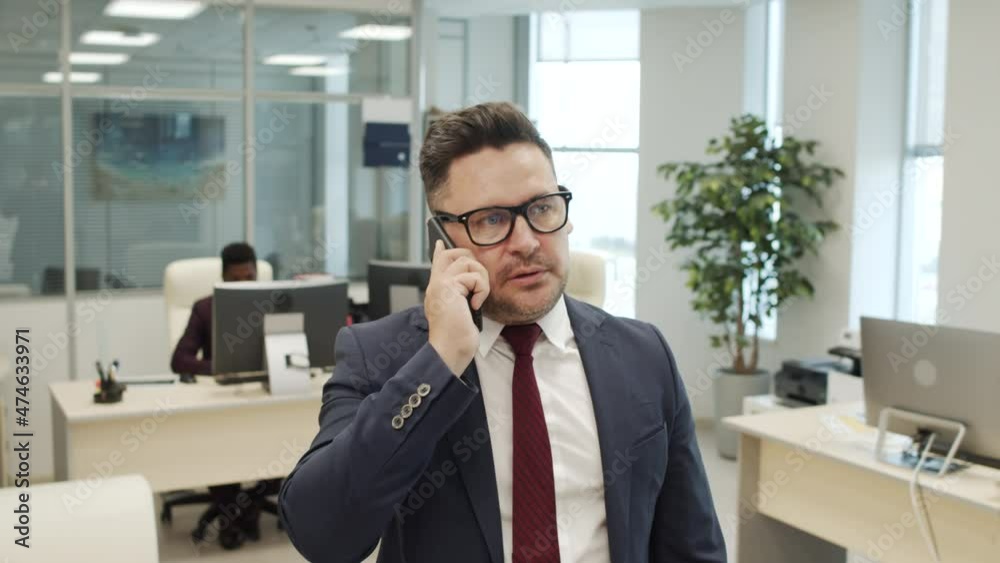 Handheld tracking shot of male executive in suit and glasses pacing in office and talking on mobile phone