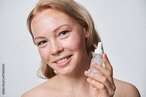 Smiling freckled woman holding bottle with serum for skin care