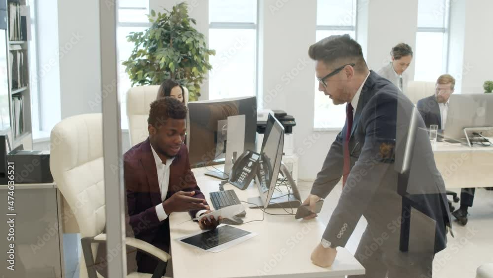 Medium shot through glass wall in office Caucasian male CEO in suit and necktie standing by desk of African-American male corporate manager and discussing document Medium shot through glass wall in office Caucasian male CEO in suit and necktie standing by desk of African-American male corporate manager and discussing document