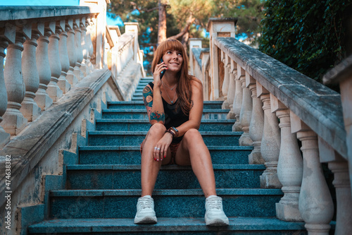 A young smiling woman with tattoos is sitting on the stome stairs and talking on the phone. Bottom view. Communication and business concept photo