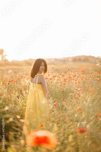 Fototapeta Naklejka Na Ścianę i Meble -  Beautiful young woman in a yellow dress walking in a poppy field on a summer day. Girl enjoying flowers in the countryside. Selective focus