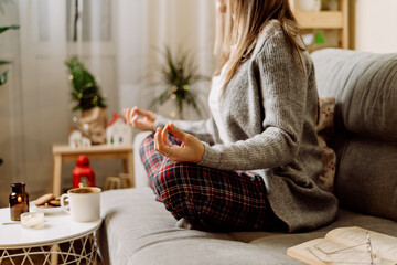 Cozy woman in knitted winter warm sweater and checkered pajama drinking hot cocoa or coffee in mug, reading book, meditation yoga on couch at home. Christmas holidays with decor and lights