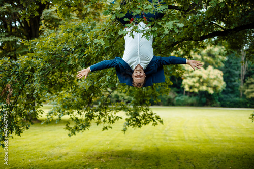 Playful businessman with arms outstretched hanging upside down from tree at park
