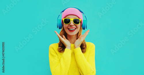 Portrait of happy surprised smiling young woman model with headphones listening to music wearing colorful yellow sweater on blue background