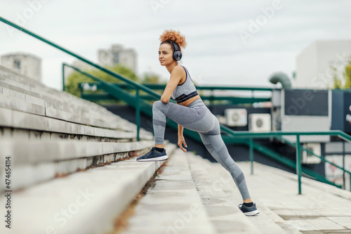 A slim sportswoman with curly hair listening to music on headphones and stretching her legs while standing on the stairs in an urban part of the city. Urban sportswoman training outdoors