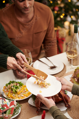 Woman using two wooden spatulas when putting portion of tasty salad in plate of guest at Christmas dinner