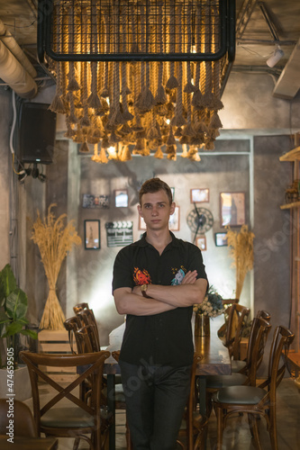 Portrait of young caucasian man in black polo standing in cafe with crossed hands and looking at camera with copy space. High quality photo
