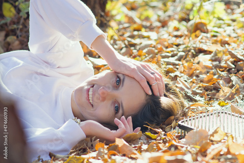 Happy young woman lies next to sadhu yoga board on the ground with autumn foliage in the park. A girl in white clothes smiles happily  looks at the camera  lying on the ground in the forest. Copyspace
