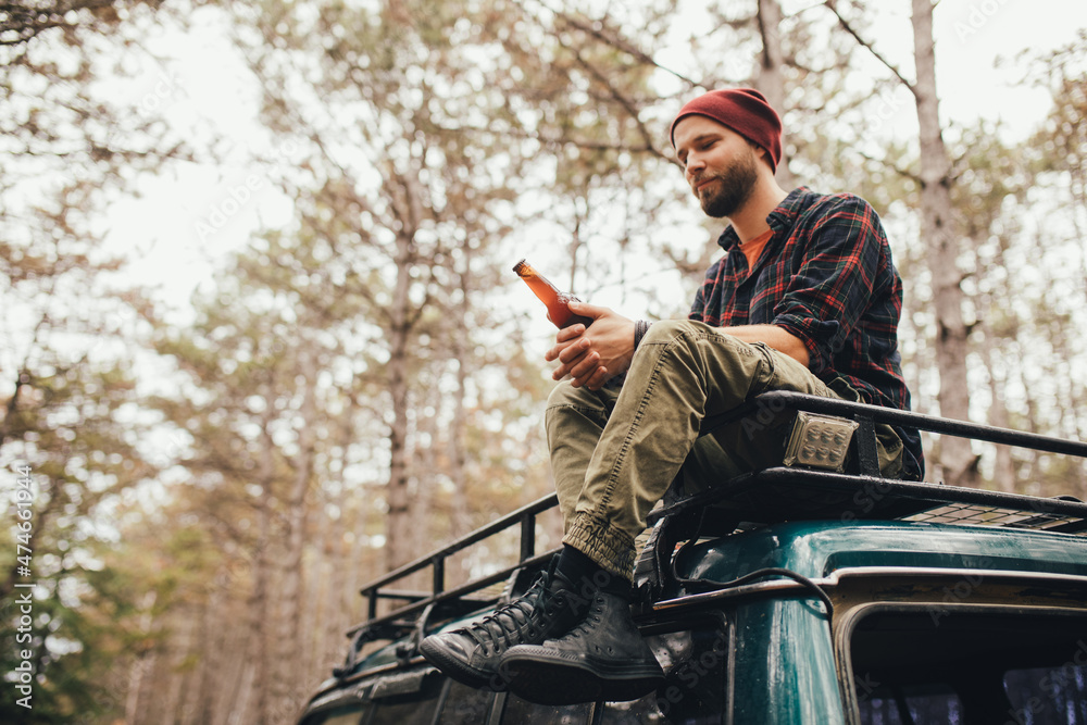 Millennial man hipster with beard sitting on car roof drinking beer in a pine forest.