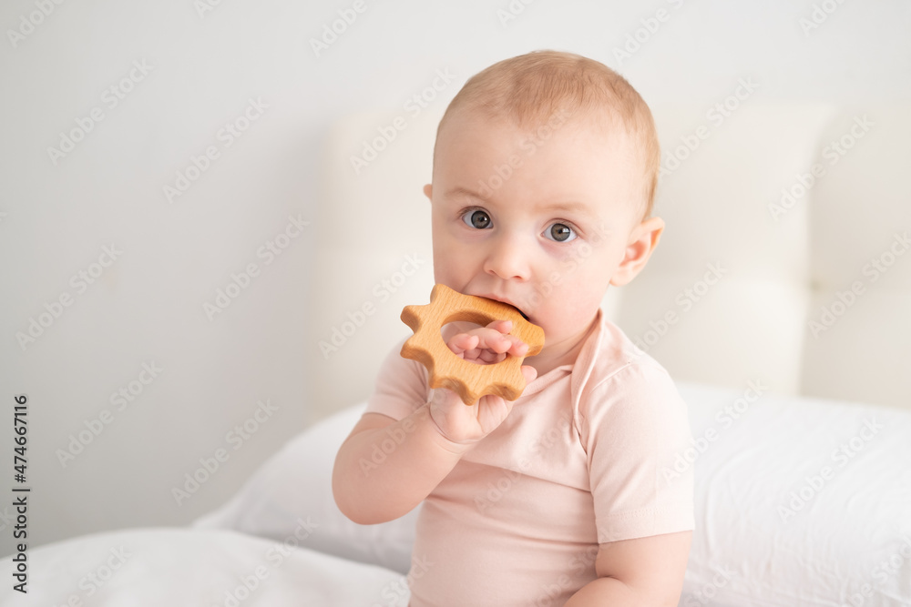 baby girl in light pink bodysuit nibbling wooden toys on white bedding on bed baby girl in light pink bodysuit nibbling wooden toys on white bedding on bed
