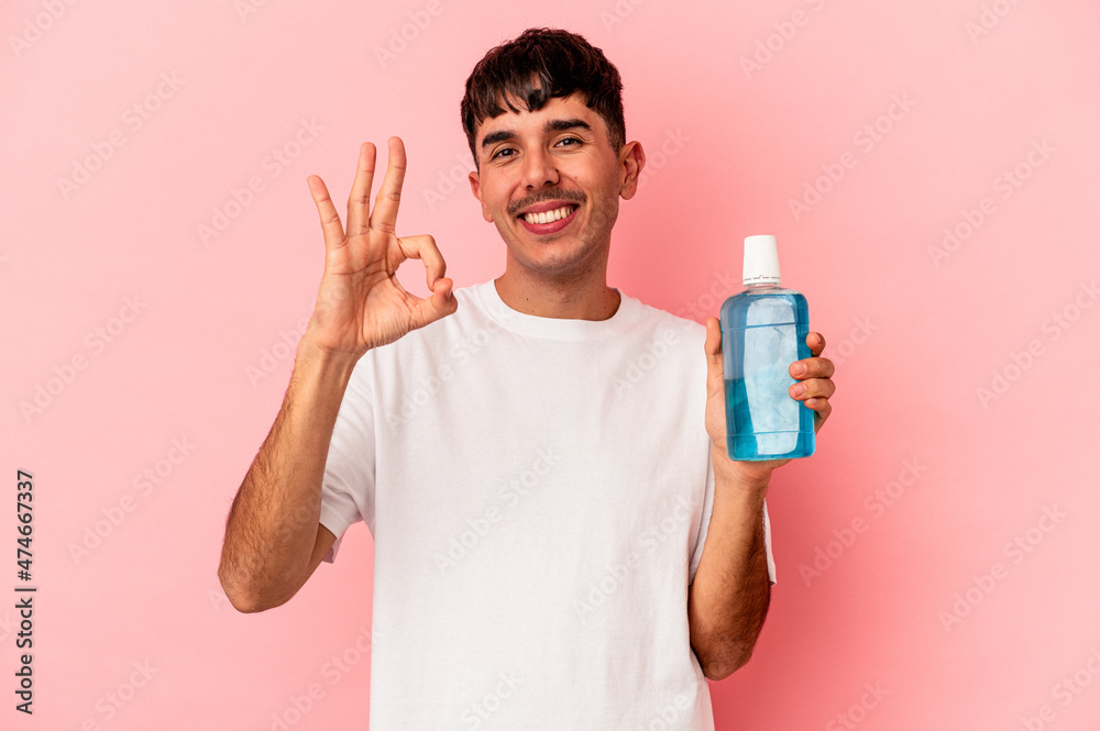 Young mixed race man holding mouthwash isolated on pink background cheerful and confident showing ok gesture. Young mixed race man holding mouthwash isolated on pink background cheerful and confident showing ok gesture.