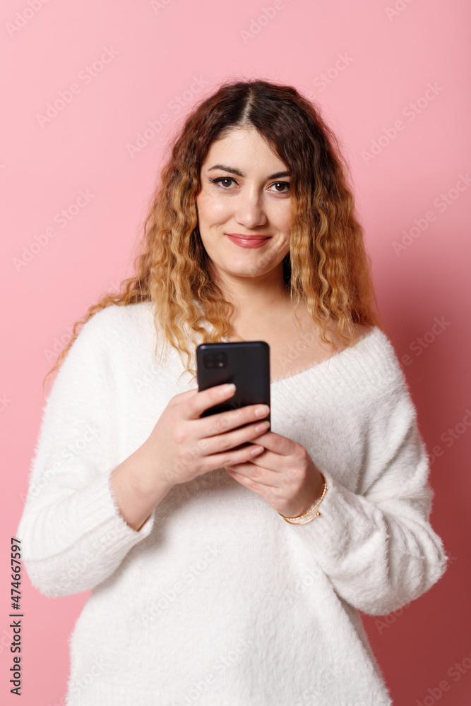 A cheerful young woman, standing isolated on a pink background with a smartphone in her hands, looks into the camera and smiles. A cheerful young woman, standing isolated on a pink background with a smartphone in her hands, looks into the camera and smiles.