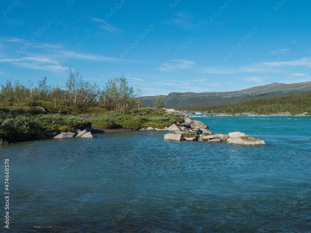 View of turquoise blue water of wild Vuojatadno river. Northern landscape in Swedish Lapland with birch tree forest and green mountains at Padjelantaleden hiking trail. Summer sunny day, blue sky
