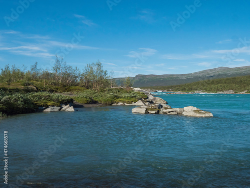 View of turquoise blue water of wild Vuojatadno river. Northern landscape in Swedish Lapland with birch tree forest and green mountains at Padjelantaleden hiking trail. Summer sunny day, blue sky