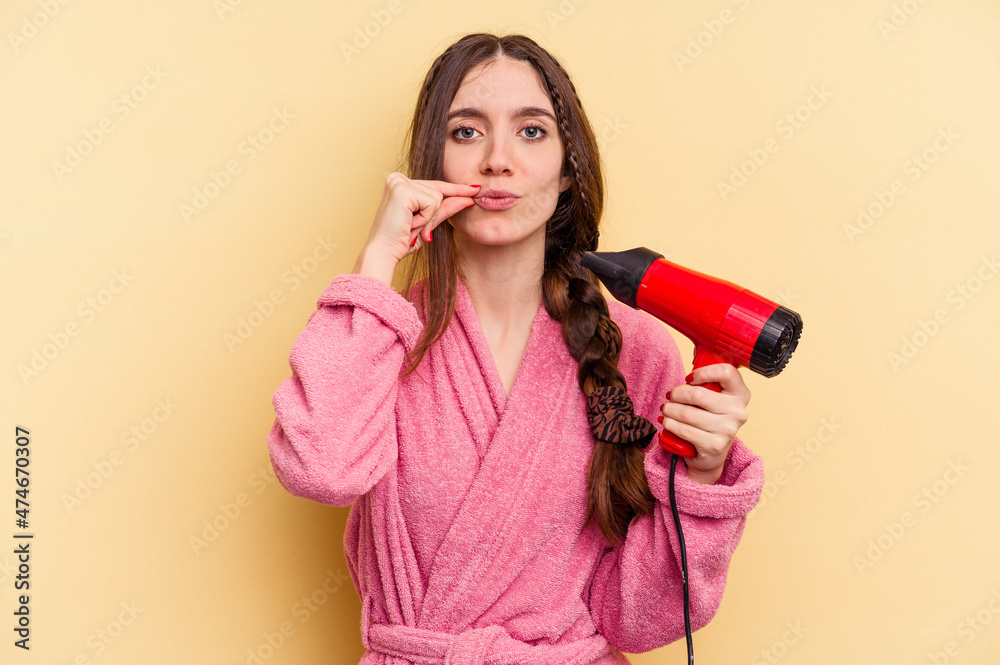Young woman holding a hairdryer isolated on yellow background with fingers on lips keeping a secret. Young woman holding a hairdryer isolated on yellow background with fingers on lips keeping a secret.