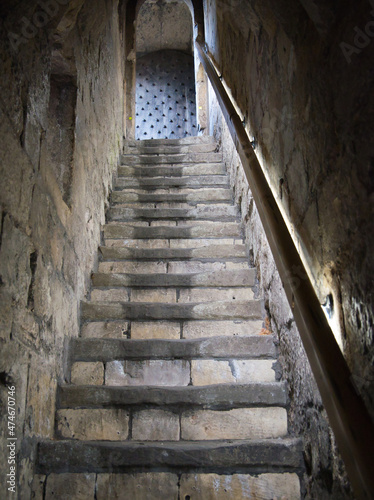 A steep flight of very old, stone steps between two stone walls leading to a studded wooden door. A handrail runs the length of the stairs on the right side.
