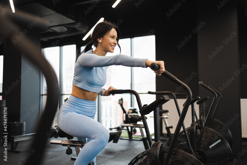 Fototapeta premium Cheerful woman riding on exercise bike. Cardio workout in the gym