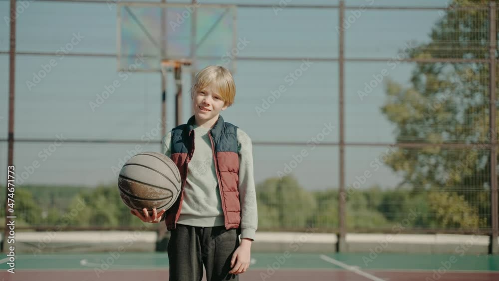 Boy stands on a court holding basketball ball and looking at the camera. Boy training street basketball. Focused streetball player practicing basketball alone outdoor. High quality 4k footage Boy stands on a court holding basketball ball and looking at the camera. Boy training street basketball. Focused streetball player practicing basketball alone outdoor. High quality 4k footage