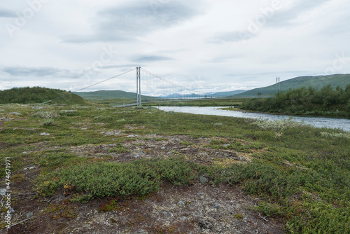 Northern landscape in Swedish Lapland with pedestrian suspension bridge over Vuojatadno river, birch tree forest and green mountains. Summer overcast day, moody sky photo