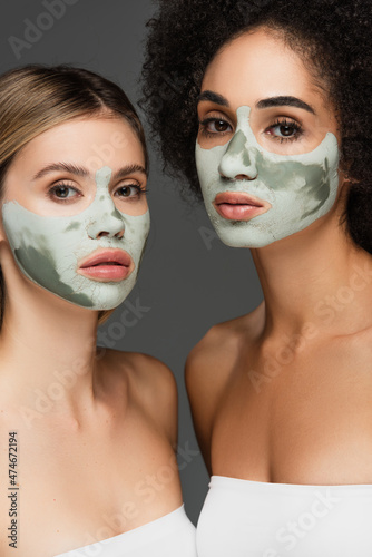 young multiethnic women looking at camera while pampering faces with clay masks isolated on grey
