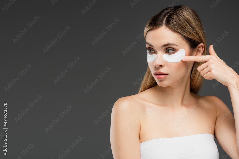 young woman looking away while pointing at eye patch on face isolated on grey young woman looking away while pointing at eye patch on face isolated on grey