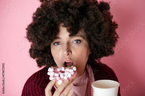 Woman with Afro hairstyle, Taken bite Of donut with marshmallow and holding in hand paper cup with coffee.