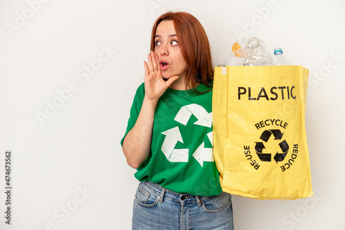 Young caucasian woman recycled plastic isolated on white background is saying a secret hot braking news and looking aside