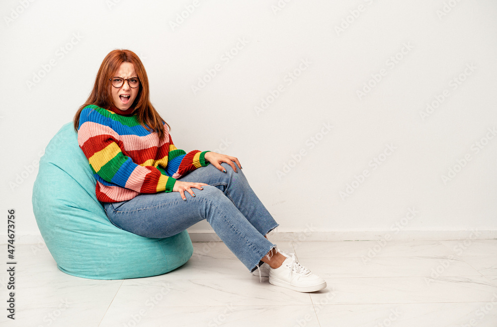 Fototapeta premium Young caucasian woman sitting on puff isolated on white background screaming very angry and aggressive.