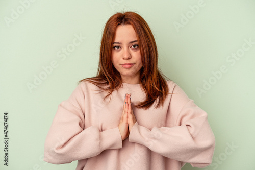 Young caucasian woman isolated on green background praying, showing devotion, religious person looking for divine inspiration. © Asier