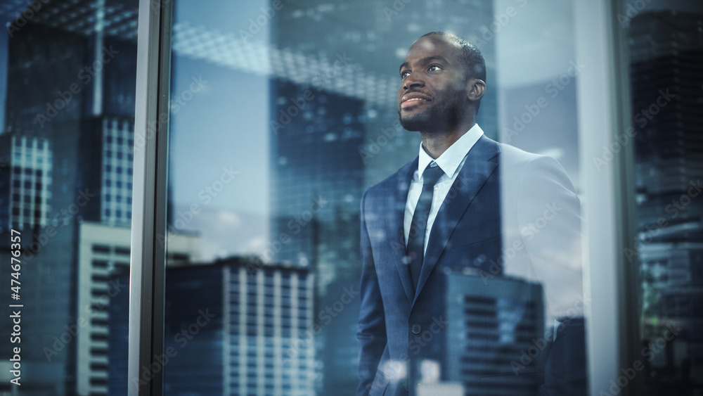 Fototapeta premium Thoughtful African-American Businessman in a Perfect Tailored Suit Standing in His Office Looking out of the Window on Big City. Successful Investment Manager Planning Strategy. Outside Shot