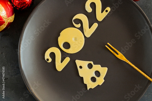 Figures 2022 and furtree made of cheese with holes, on a black plate with small fork and Christmas tree decoration, symbol of proper nutrition, menu. Horizontal. Winter and New year concept photo