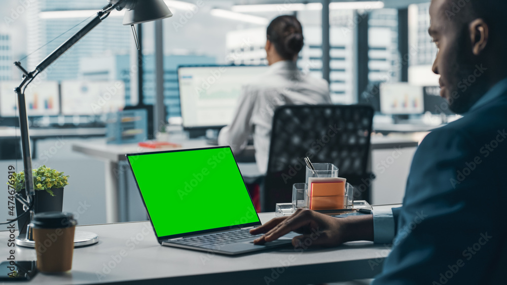 Successful Black Businessman Sitting at Desk Working on Green Screen Chroma Key Laptop Computer in Big City Office. Hard Working Top Manager Doing Research for e-Commerce Project. Over Shoulder