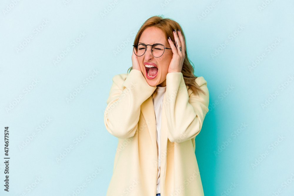 Young caucasian woman isolated on blue background covering ears with hands trying not to hear too loud sound. Young caucasian woman isolated on blue background covering ears with hands trying not to hear too loud sound.