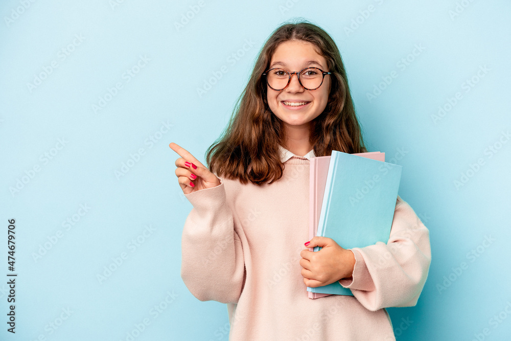 Little caucasian student girl holding books isolated on blue background smiling and pointing aside, showing something at blank space.