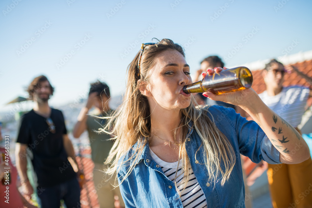 Fototapeta premium Portrait of pretty woman with fair hair at party. Young girl in striped top and shirt drinking beer. Friends of different nationalities in background. Party, friendship concept
