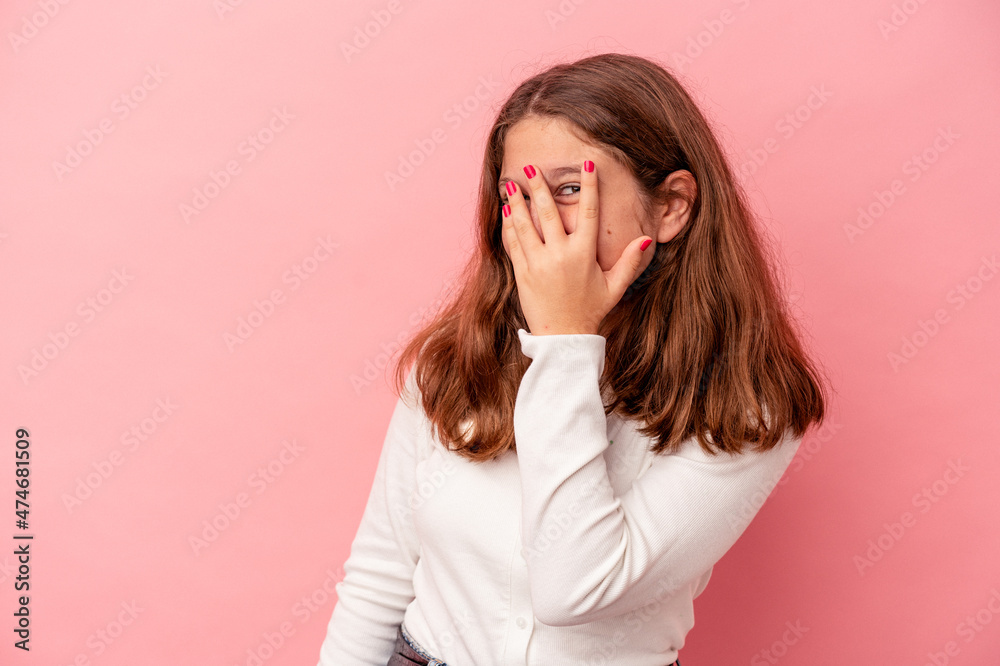 Little caucasian girl isolated on pink background blink at the camera through fingers, embarrassed covering face.